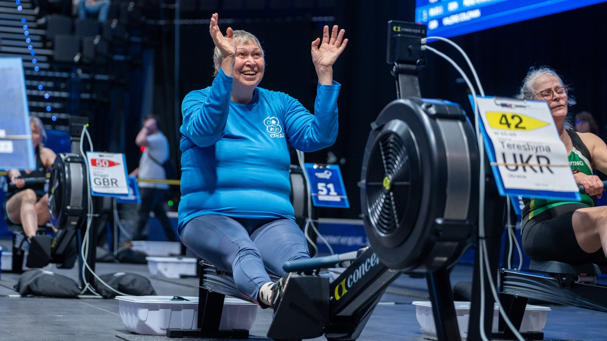 Woman waving from a RowErg at an indoor race